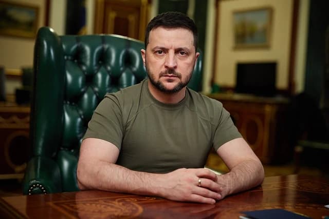 Volodymyr Zelenskyy, President of Ukraine, sits at a desk with his hands clasped.