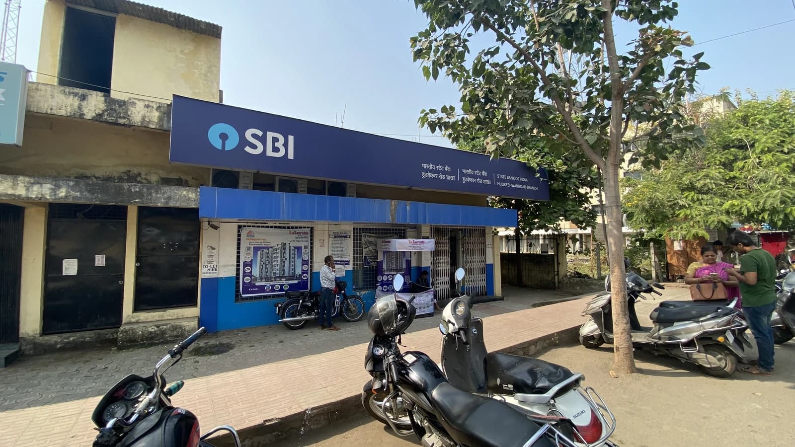 Exterior of a State Bank of India branch with parked motorbikes and people nearby.