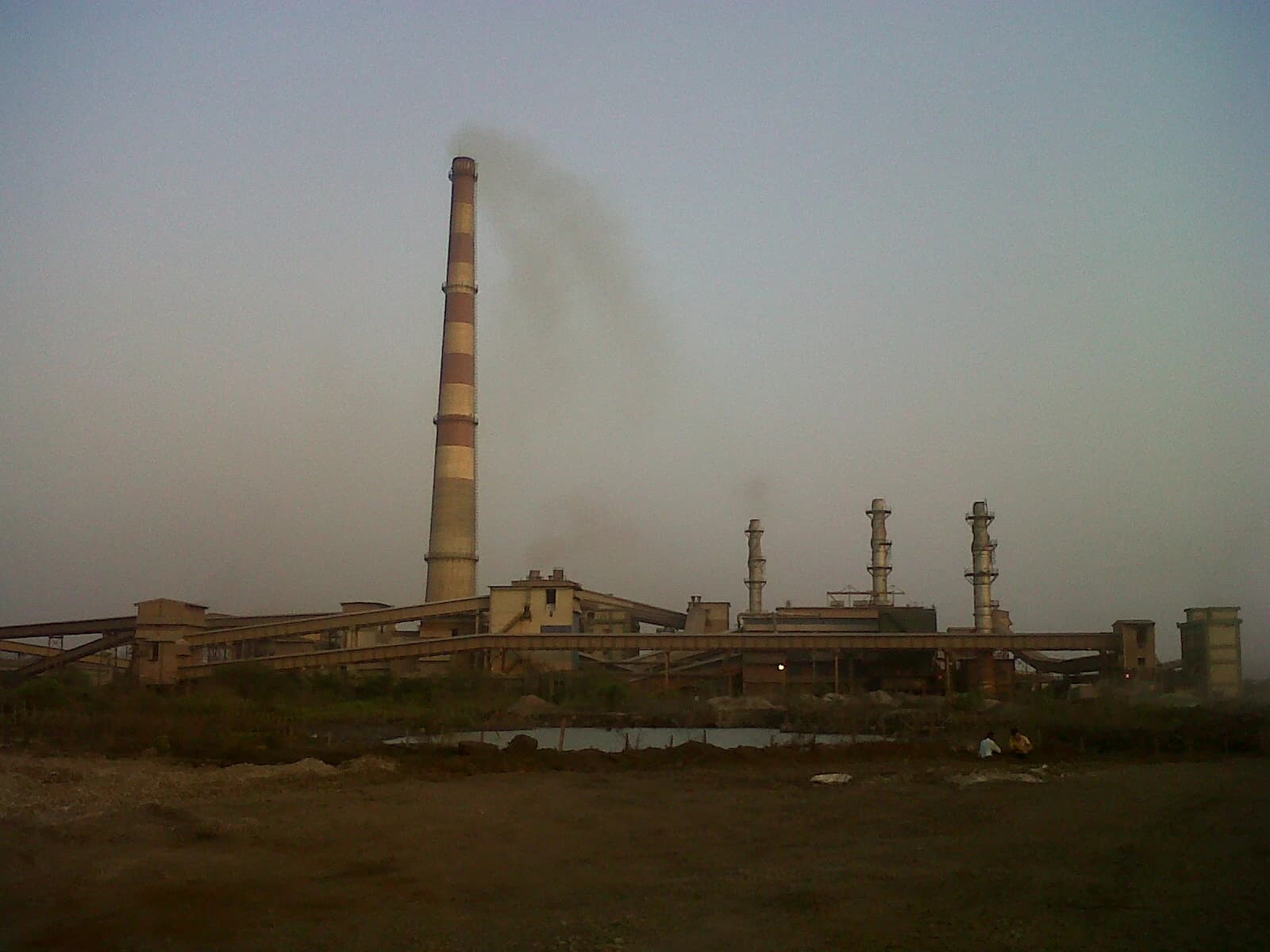 Industrial plant with tall smokestack emitting smoke, surrounded by low-lying buildings.