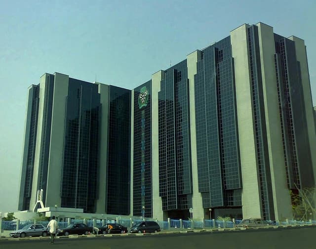 Exterior of the Central Bank of Nigeria building in Abuja, with a street and cars in the foreground.