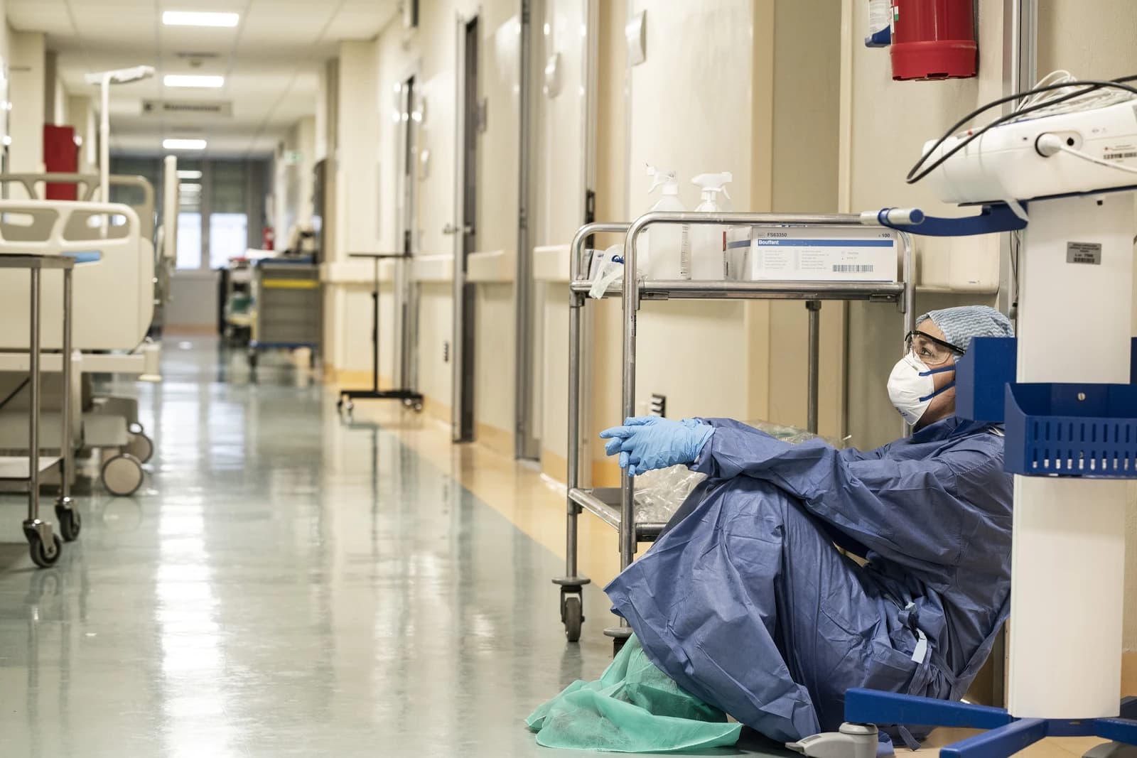 Healthcare worker, wearing scrubs and a mask, sits on a hospital corridor floor.