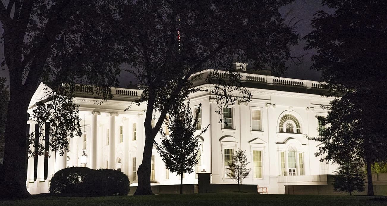 The White House illuminated at night, viewed through silhouetted trees.