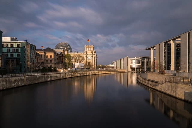 Reichstag Building and government offices reflected in Spree River under cloudy sky.