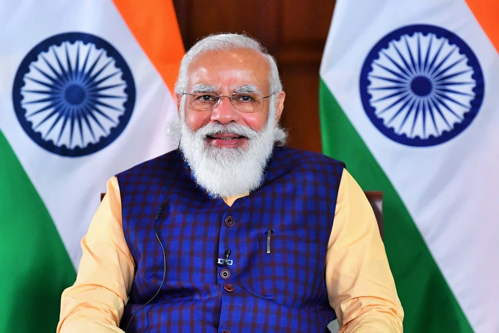 Narendra Modi, Prime Minister of India, sits smiling in front of two Indian flags.