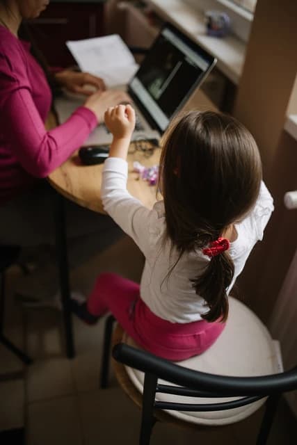 Parent working on a laptop at a table, a child playing next to them.