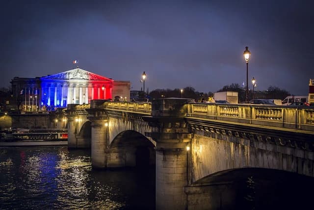 French National Assembly building illuminated in blue, white, and red by the Seine River at night.