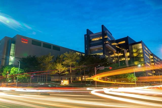 Exterior view of the TSMC Fab 12A building at dusk, with light trails from vehicles on the road.