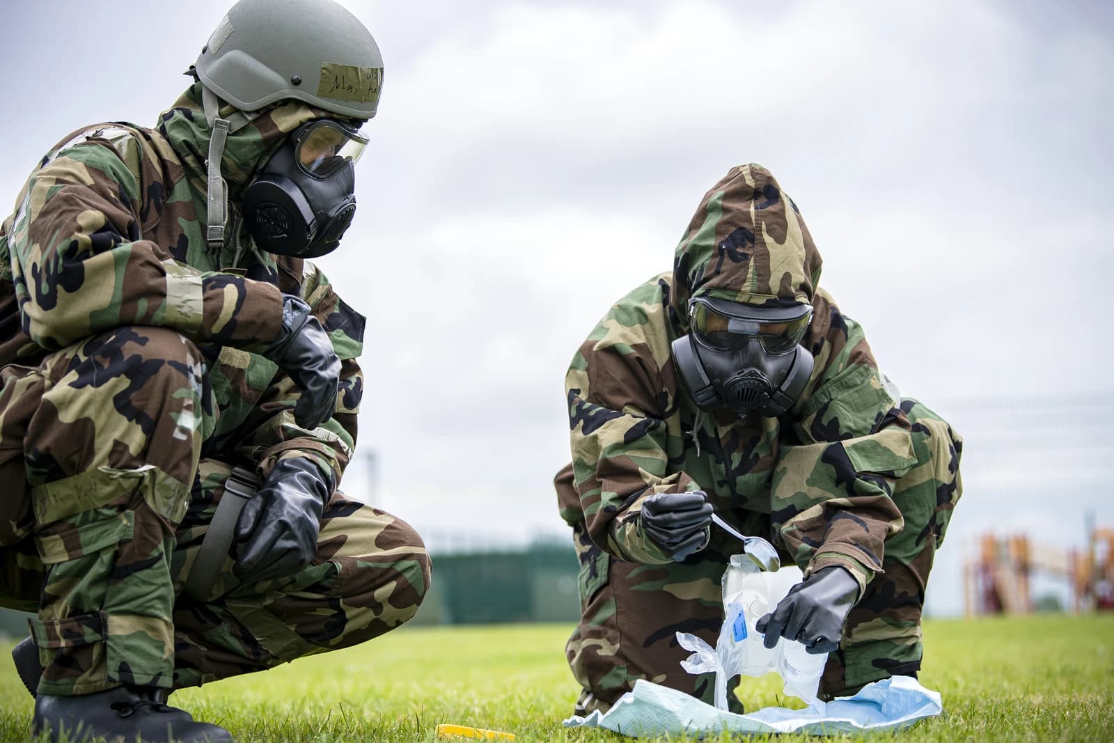 Military personnel in camouflage hazmat suits and gas masks conducting an exercise outdoors.