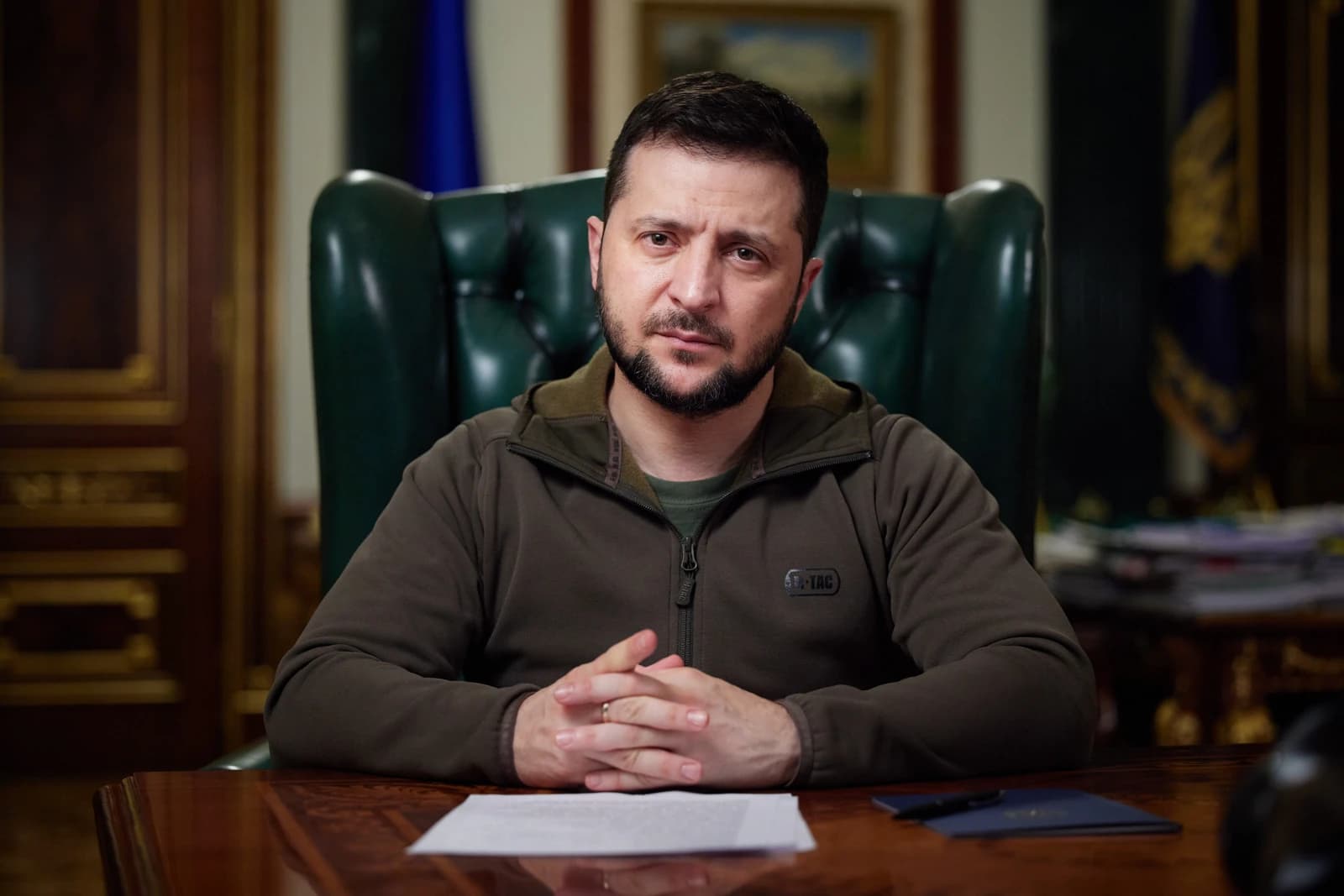 Volodymyr Zelenskyy, President of Ukraine, sits at a wooden desk with his hands clasped.