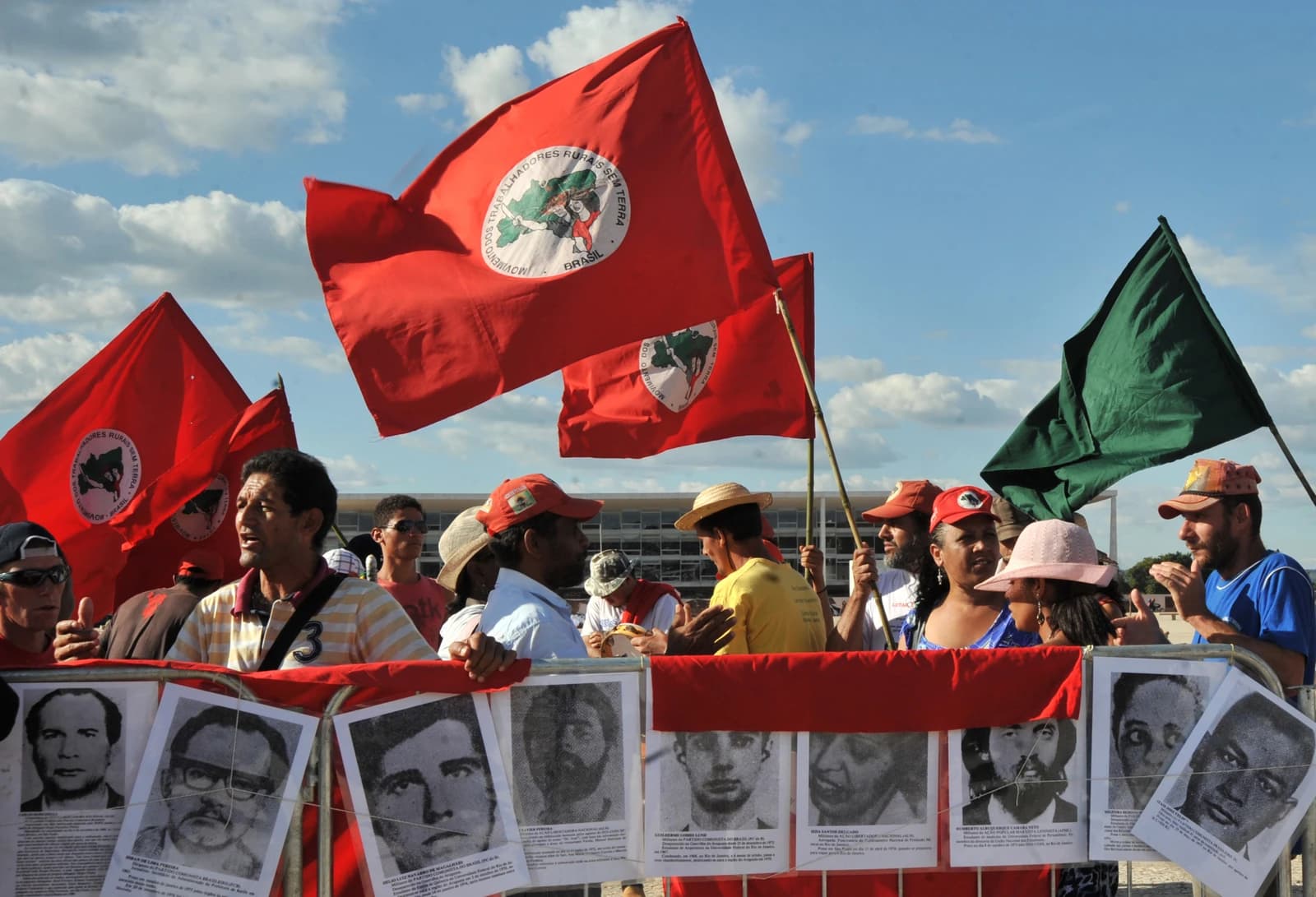 Protesters holding red flags and signs with faces of individuals, in front of a building.