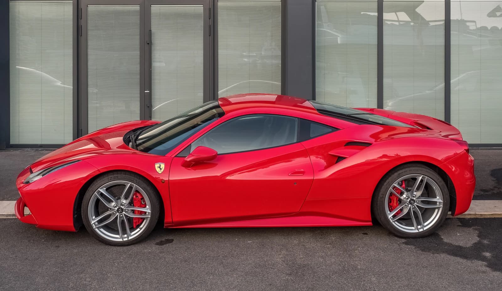 A red Ferrari parked outdoors against a building with horizontal window blinds.