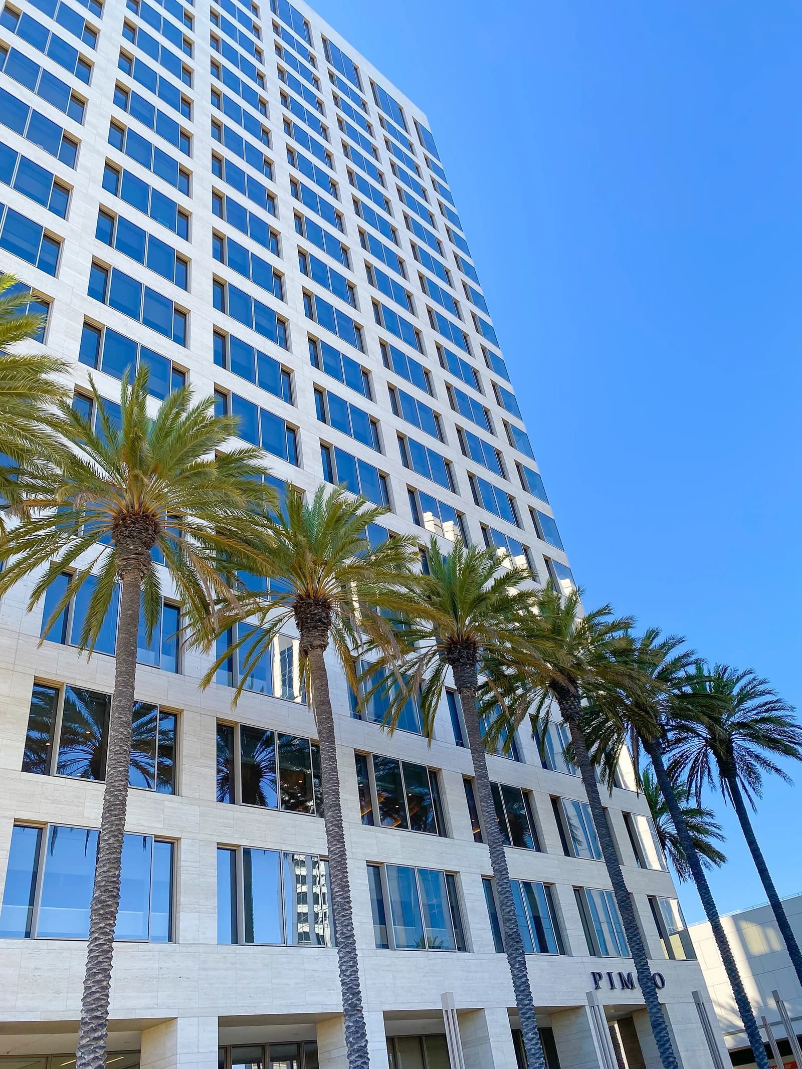 Exterior view of the Pimco headquarters building with palm trees and a clear blue sky.