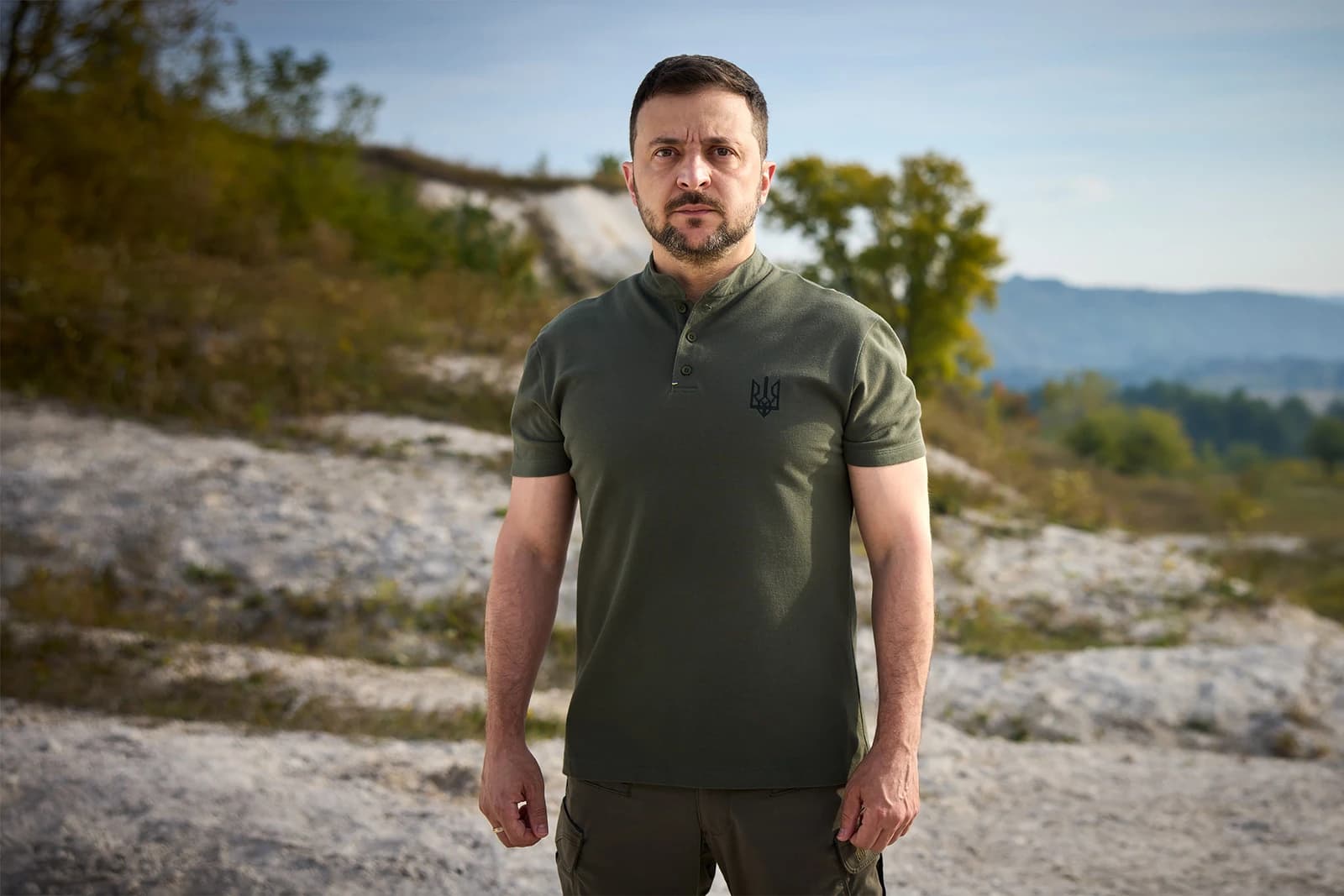 Volodymyr Zelenskyy, President of Ukraine, standing outdoors with a hilly background.