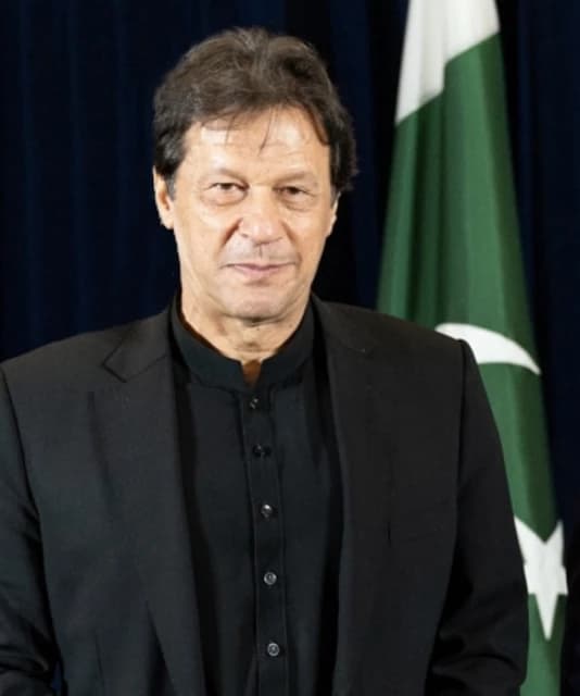 Imran Khan, former Prime Minister of Pakistan, stands with a Pakistani flag in the background.
