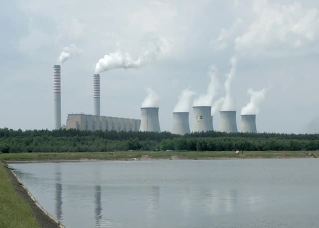 Belchatów Power Station with two tall smokestacks and six cooling towers emitting steam, viewed across a body of water.