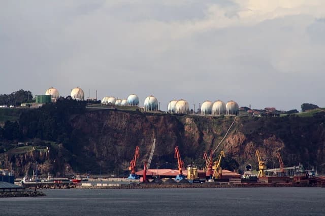 Spherical gas storage tanks visible on a hill overlooking port facilities and cranes.