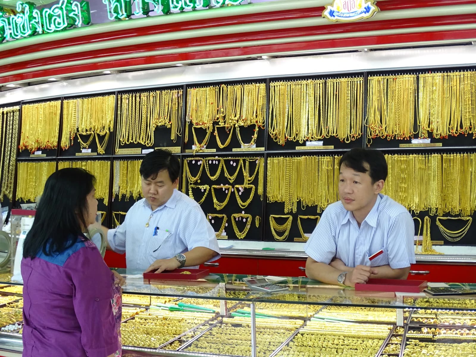 Two shop employees assist a customer at a counter filled with gold jewelry.