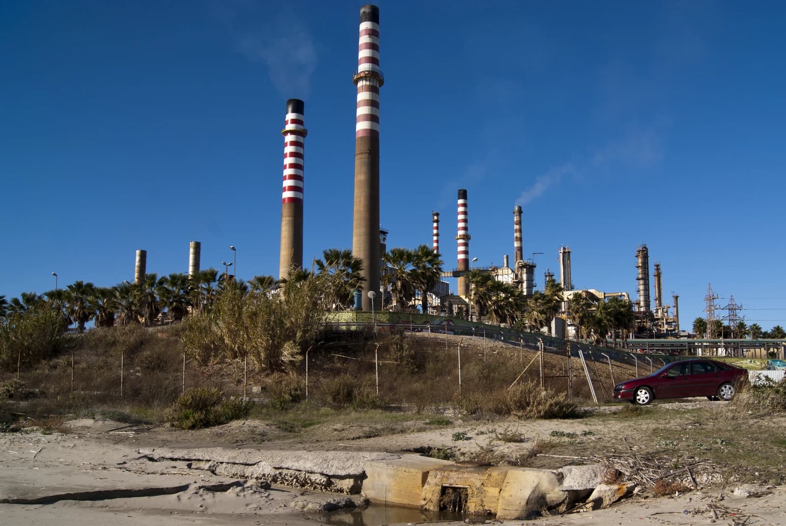 Refinery Gibraltar-San Roque showing tall industrial smokestacks and palm trees.