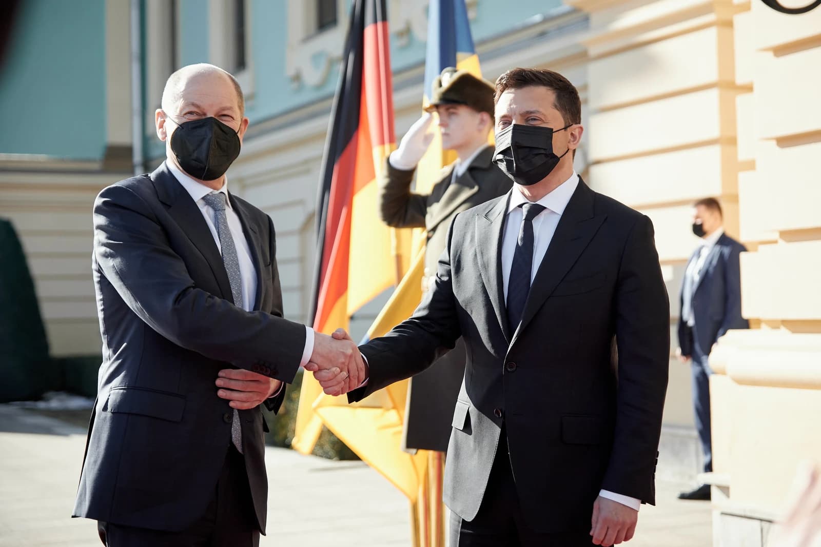 Olaf Scholz and Volodymyr Zelenskyy shaking hands outdoors, with flags in the background.