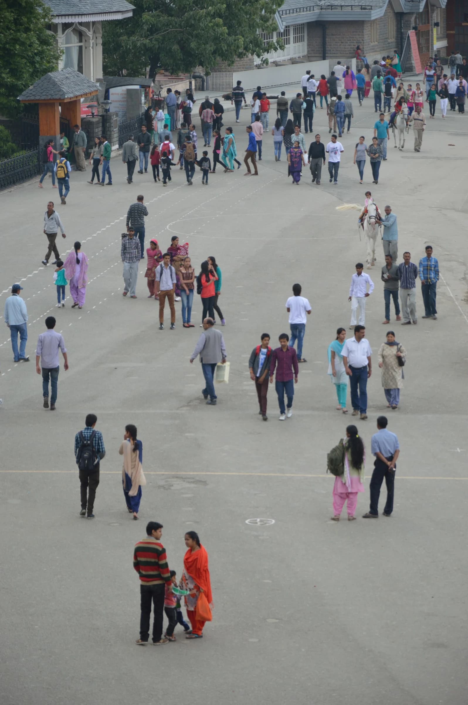 Many people, including families and individuals, walk and mill about on a wide, paved public space in a mountainous town.