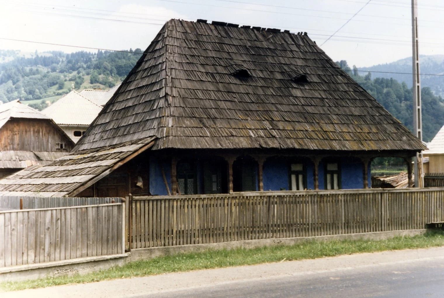 An old house with a wooden shingle roof and blue painted walls in a rural setting.