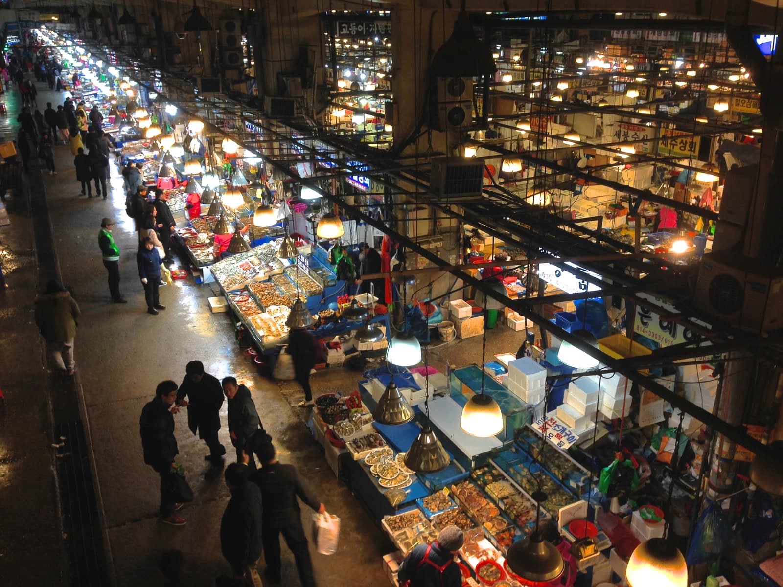 Dozens of people shop at an indoor market with many stalls selling food products.