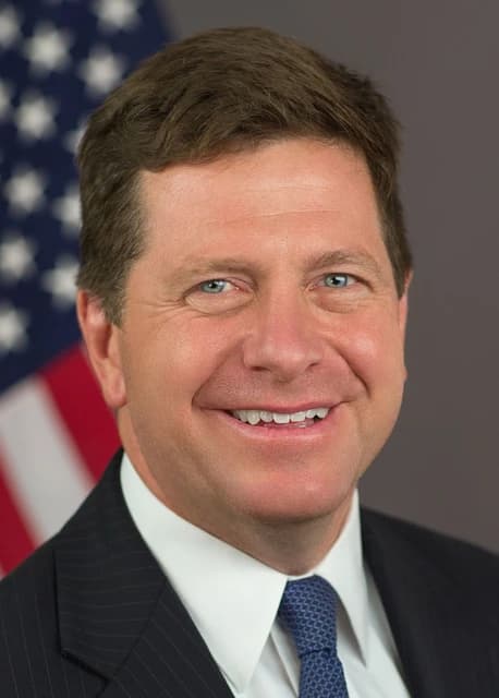 Jay Clayton, US intelligence chief, smiling for a headshot against an American flag.