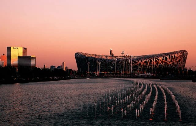Beijing National Stadium, known as the Bird's Nest, illuminated at dusk, with fountains in a foreground lake.