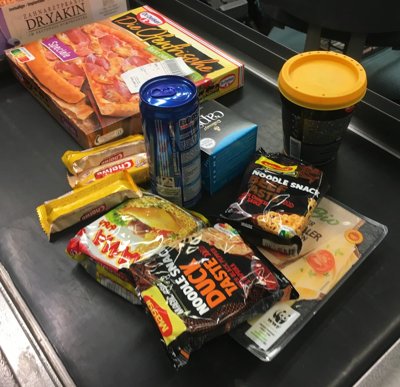 Various packaged food items like instant noodles, pizza, and cheese on a supermarket conveyor belt.