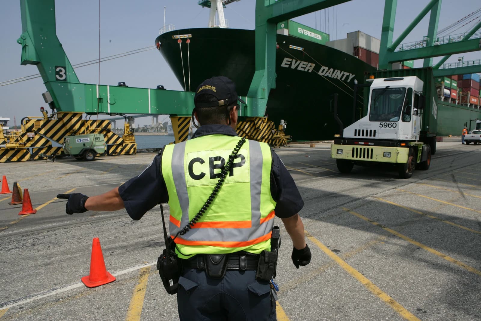 CBP officer directing a truck near a cargo ship at a port.