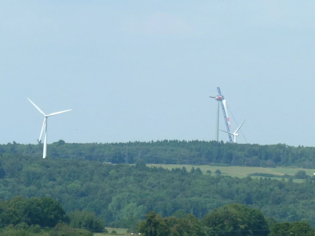 Two wind turbines standing on a hill with forested areas below them.