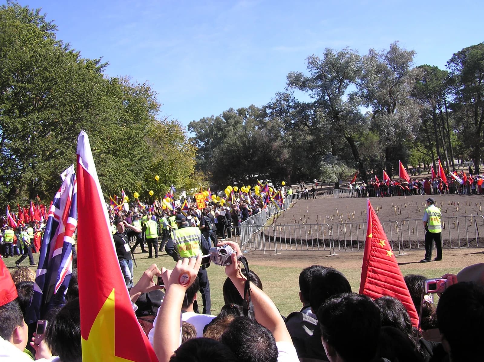 Protestors gathered outdoors, holding various flags including Tibetan and Chinese, with police observing.