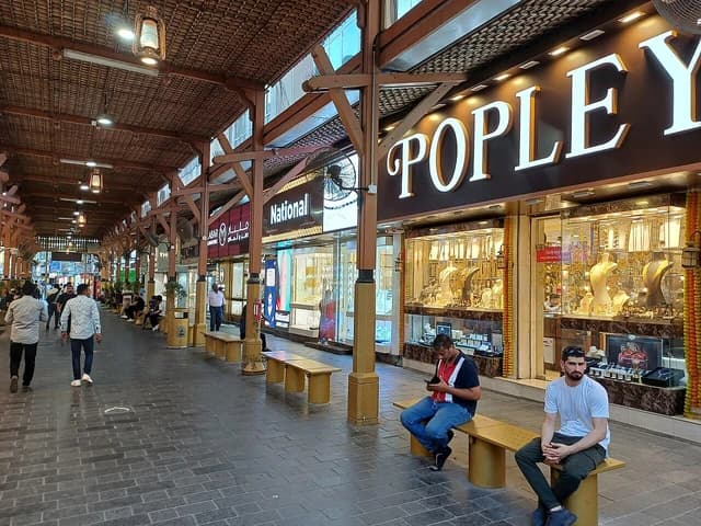 People walking through a covered marketplace, passing by shop windows displaying gold jewelry.