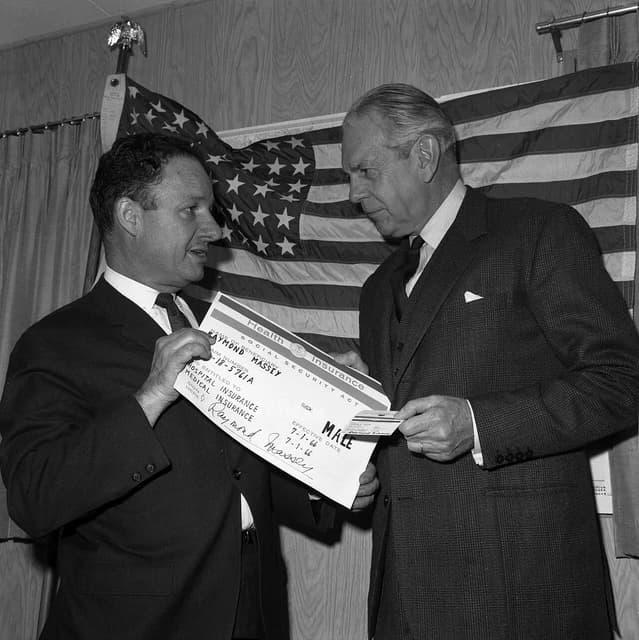 Raymond Massey, actor, receiving his Medicare identification card from an unidentified man.