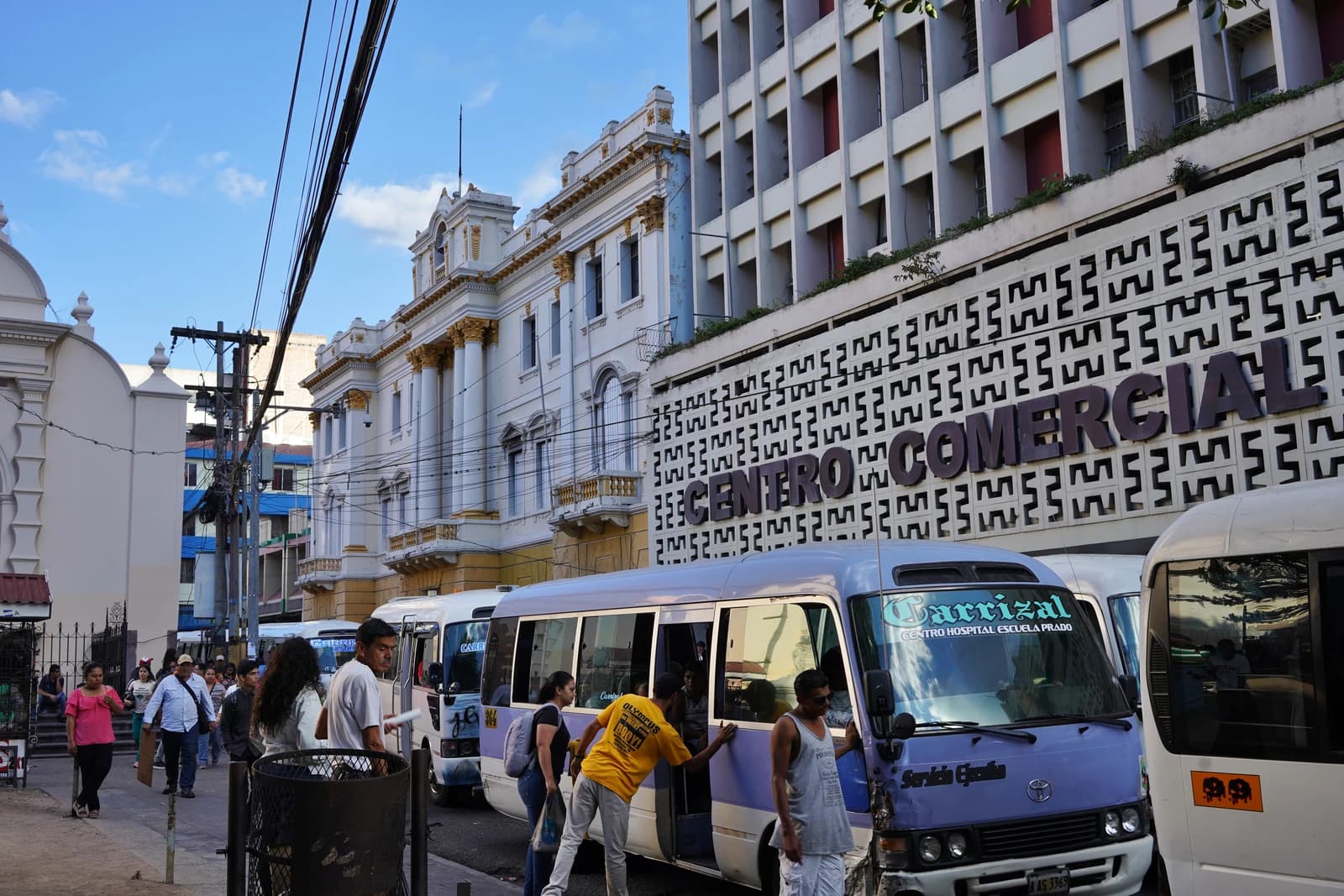 People walking on a street next to minibuses and a building with 'Centro Comercial' sign in Tegucigalpa, Honduras.