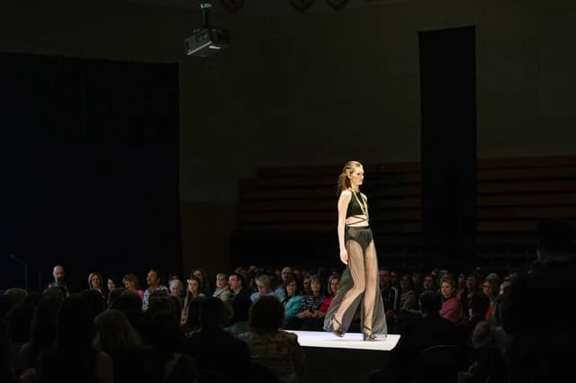 Model walks a runway during a fashion show with an audience watching.