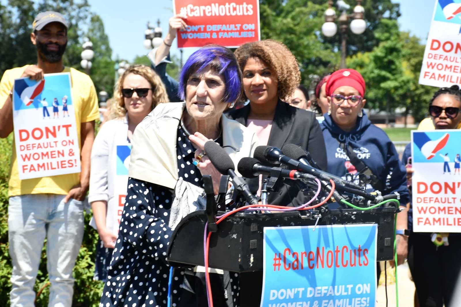 Representative Rosa DeLauro speaking at a podium with microphones, surrounded by people holding signs.