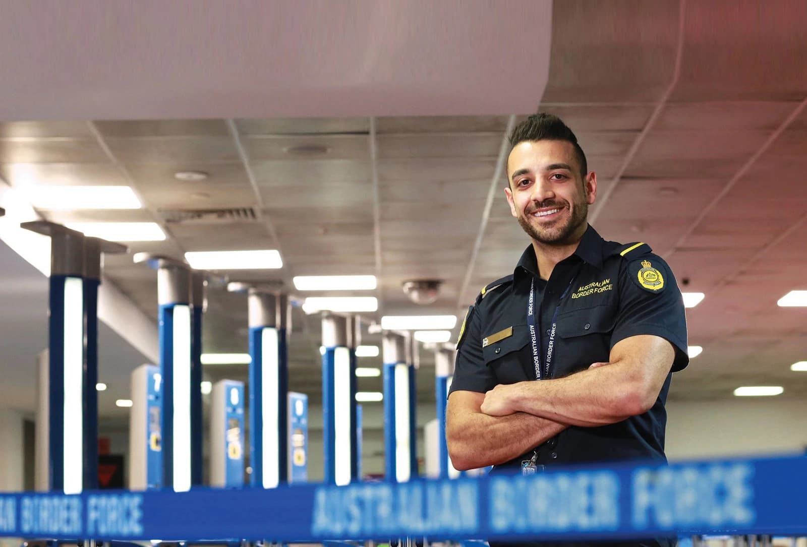 Australian Border Force officer smiling, standing with arms crossed at an airport.
