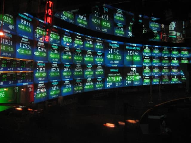 Stock market data including NASDAQ Composite and Dow Jones displayed on multiple digital screens.