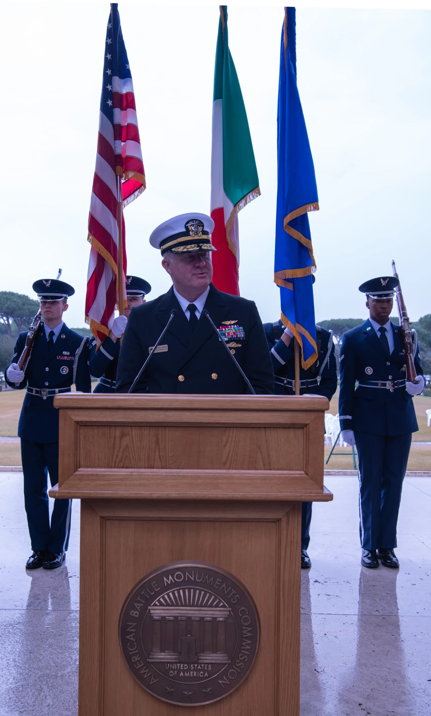 U.S. Navy Admiral speaking at a podium during an official event, with flags behind him.