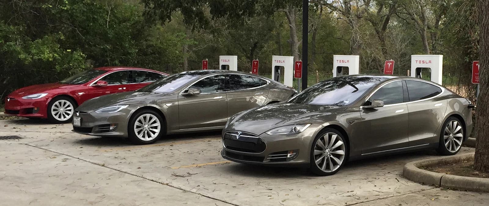 Four Tesla electric vehicles, including two gray and one red, charging at a Supercharger station.
