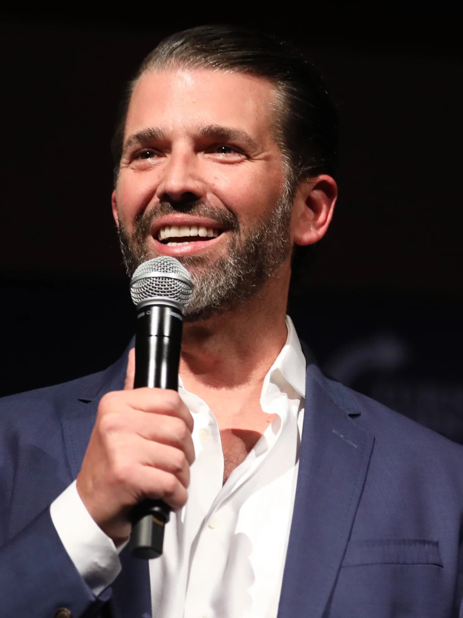 Donald Trump Jr. speaks into a microphone at an event, smiling.