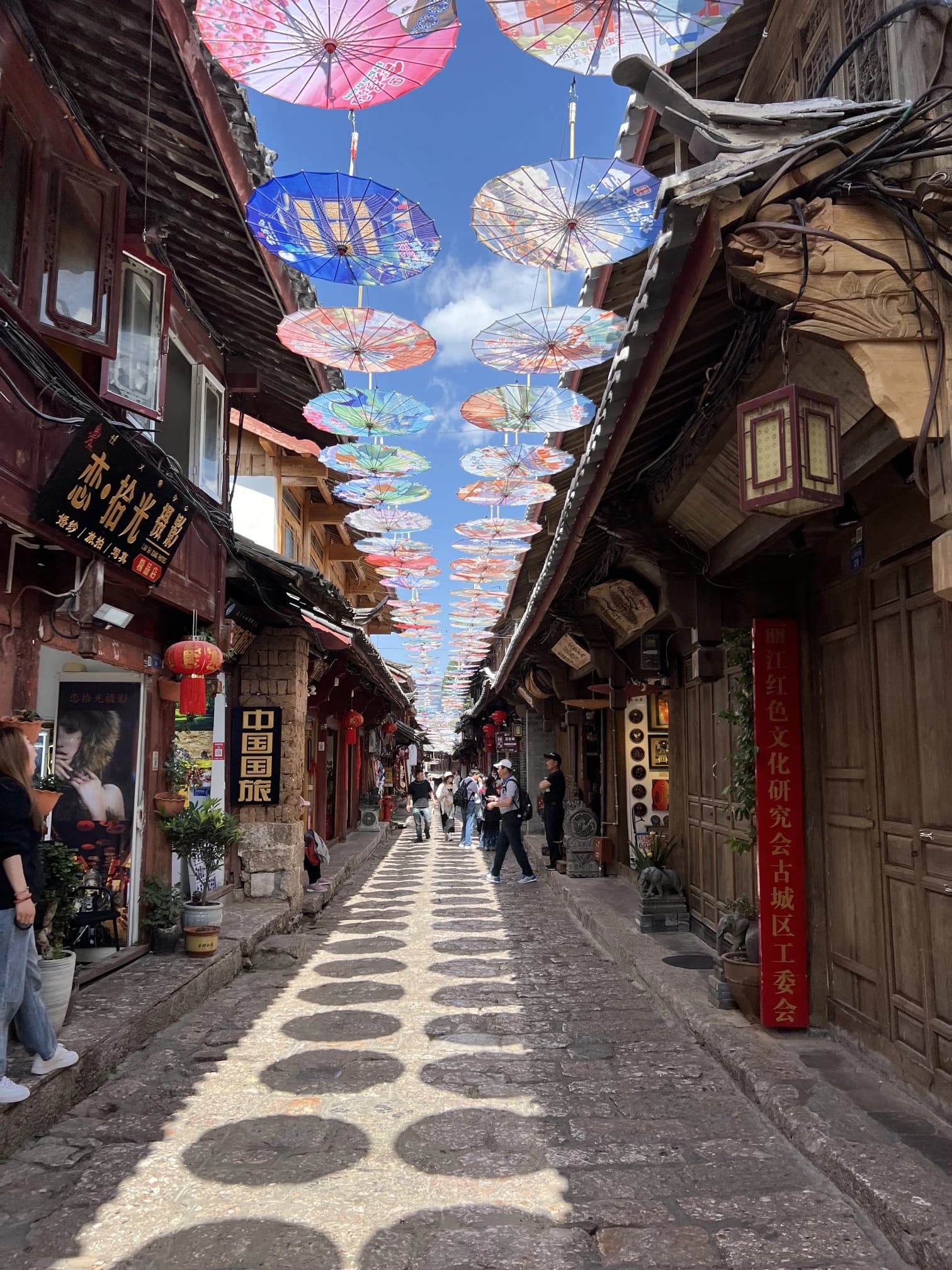 Tourists walking on a cobblestone street in Lijiang, China, with vibrant umbrellas decorating the sky.