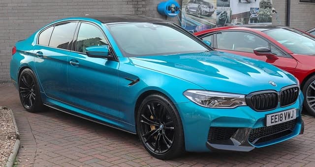 A turquoise 2018 BMW M5 sedan parked outdoors on a brick surface.