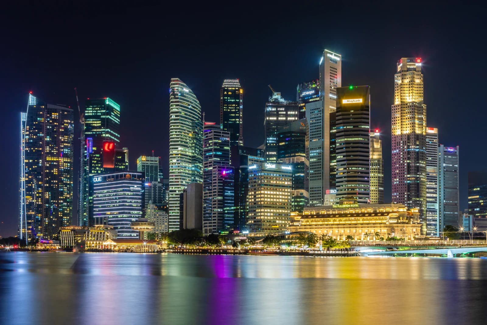 Singapore's Marina Bay skyline at night, with brightly lit skyscrapers reflecting onto the water.