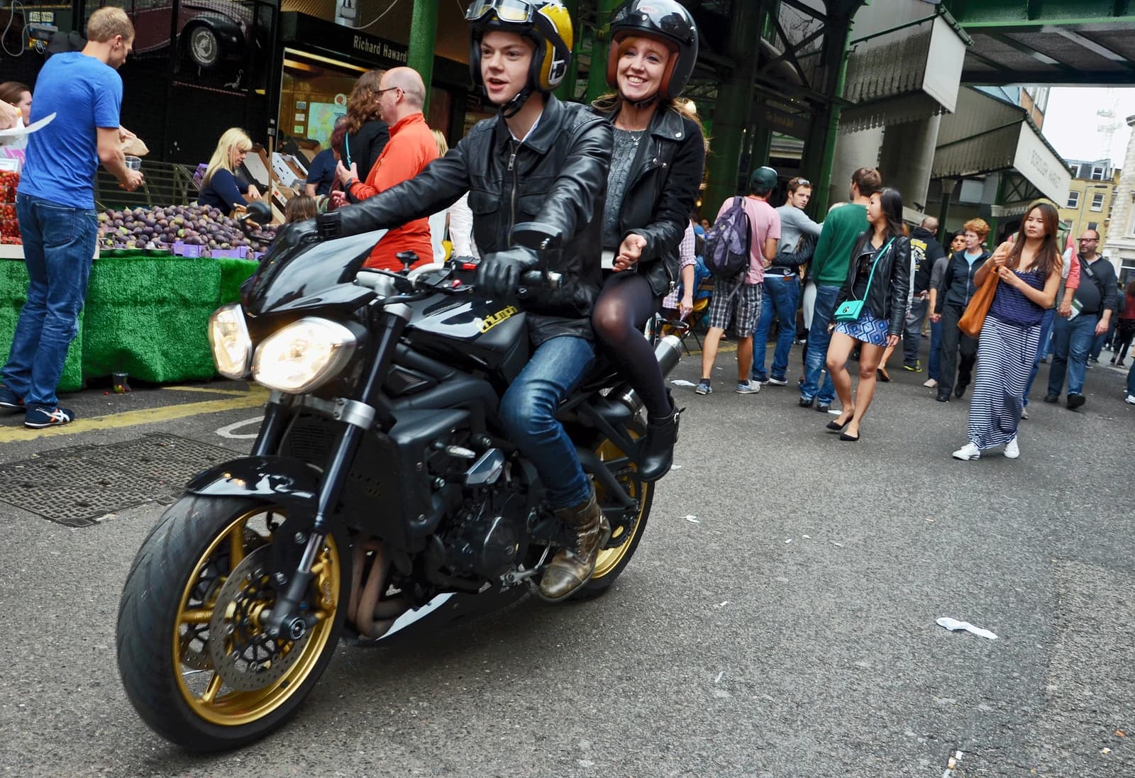 Man and woman wearing helmets riding a black motorcycle through Borough Market.