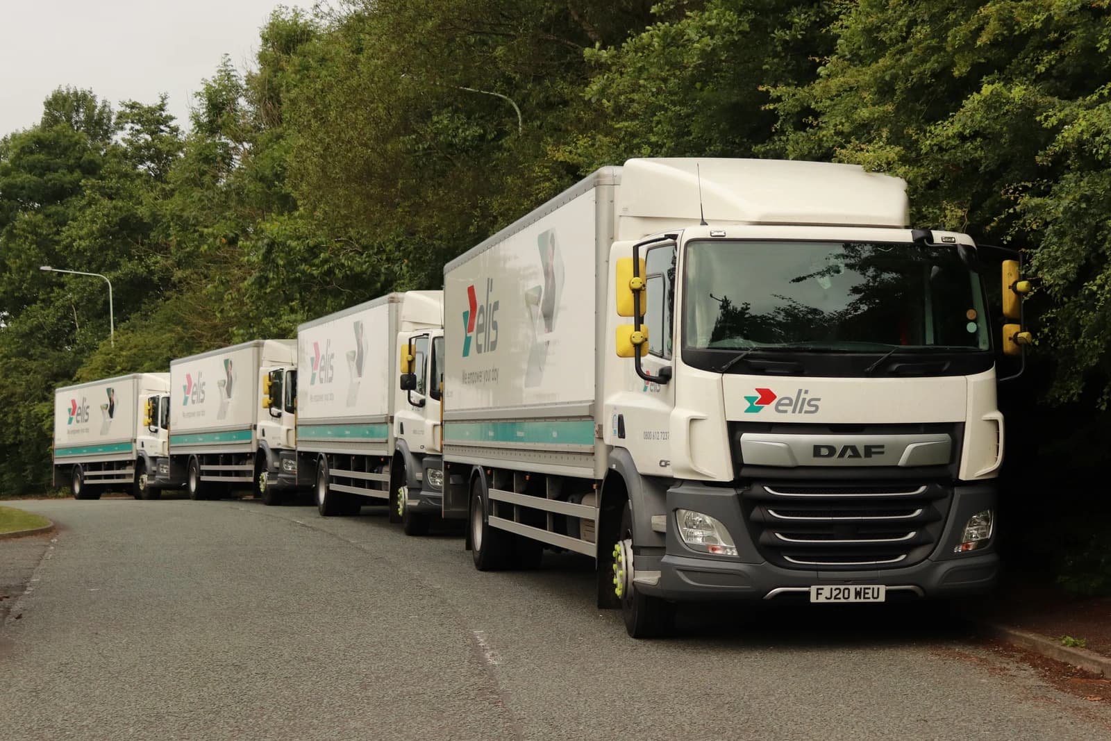 A line of white Elis trucks with DAF branding parked on an asphalt road.