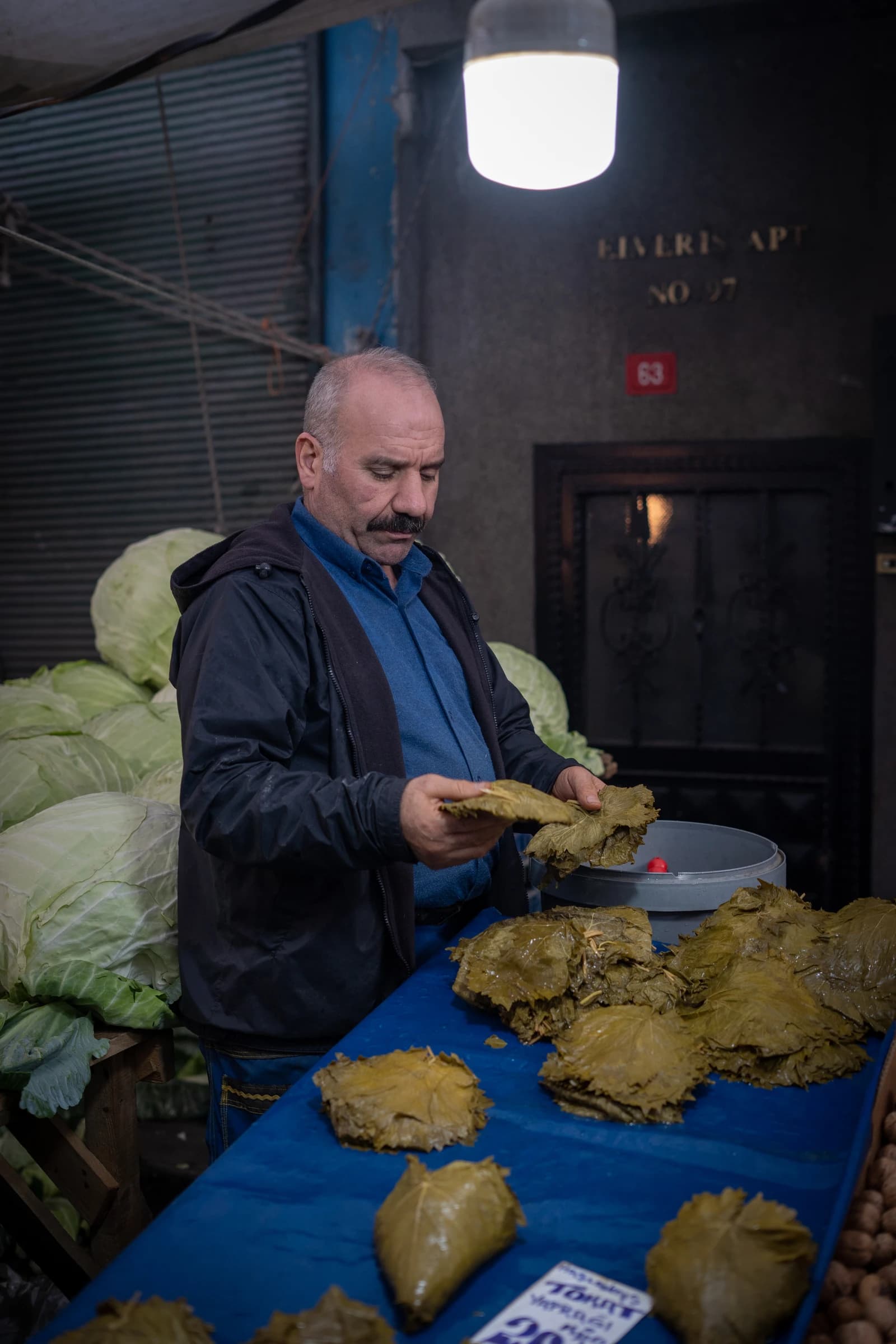 A man at a market stall arranging grape leaves on a blue cloth.