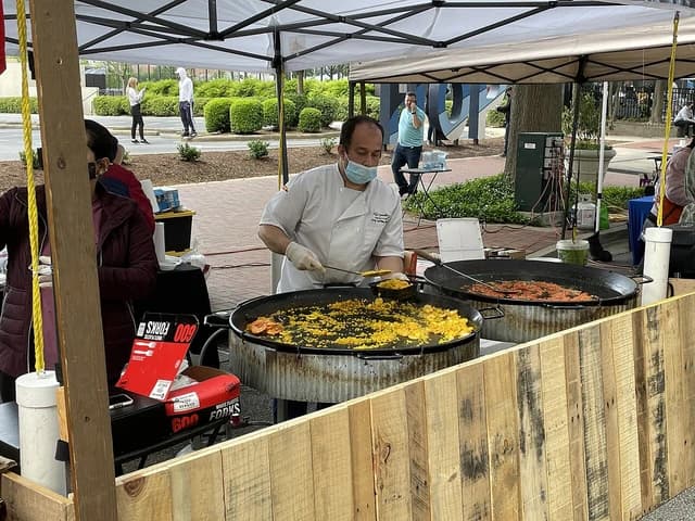 A man in a chef's coat and face mask cooking food in large pans at an outdoor market.