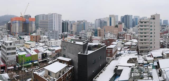 An aerial view of Seoul's urban landscape with snow on rooftops and a visible construction site.